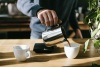 Mężczyzna trzyma w dłoni stalowy dzbanek i nalewa z niego kawę do jednej z dwóch filiżanek stojących na blacie